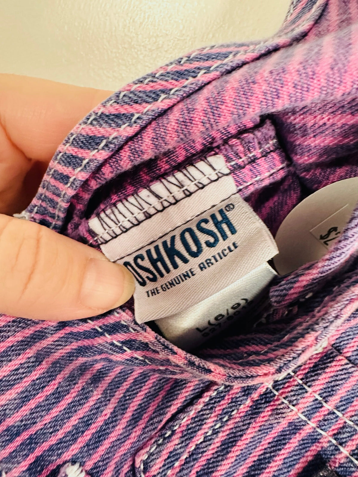 Close-up of a pink and navy striped denim shirt collar showing the 'OSHKOSH THE GENUINE ARTICLE' brand label, held by a hand.