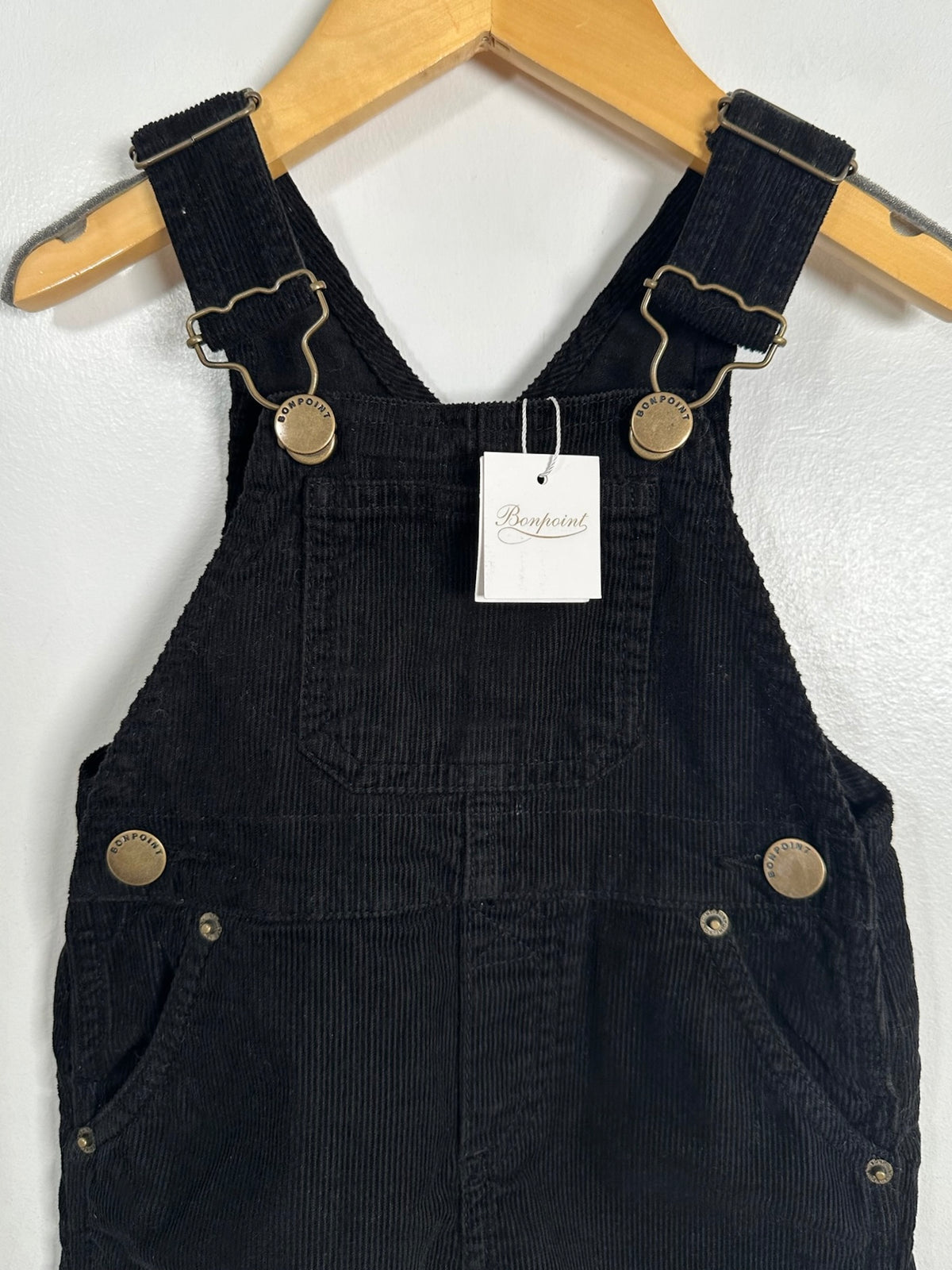 Black corduroy baby overalls hanging on a wooden hanger against a white wall, featuring adjustable straps with metal hardware and a white brand tag.