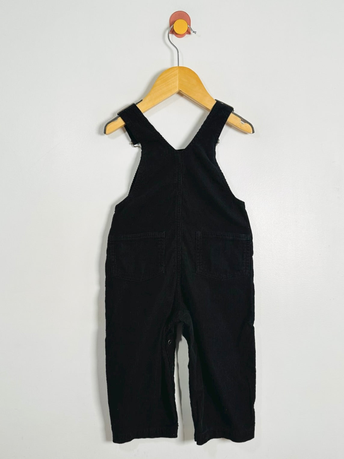 Black corduroy baby overalls hanging on a wooden hanger against a plain white wall.