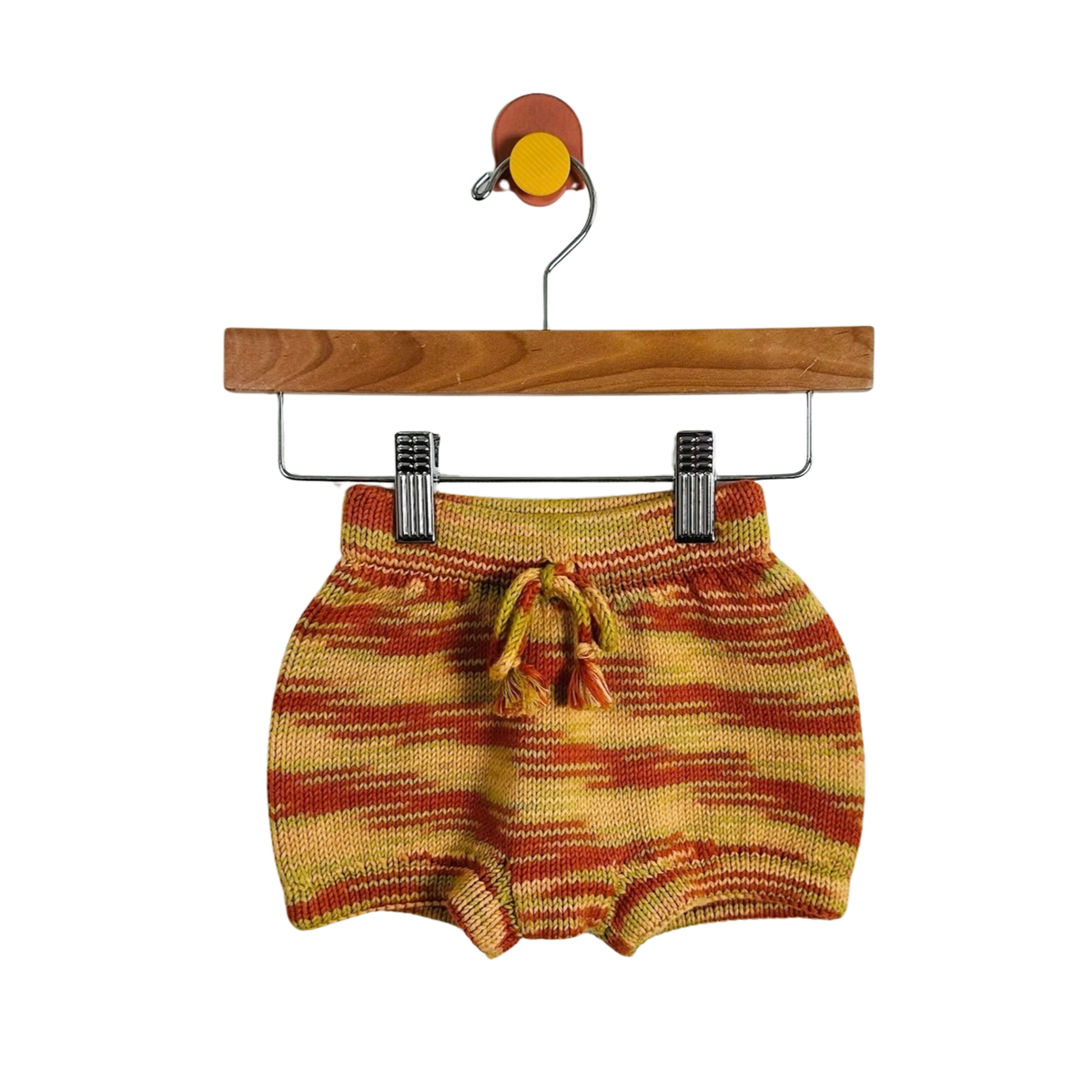 A pair of knit baby shorts in a yellow, orange, and red striped pattern, hanging from a wooden hanger with metal clips against a plain white background.