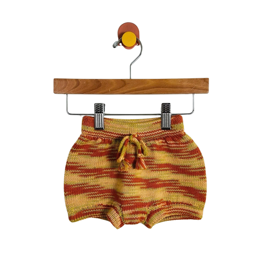 A pair of knit baby shorts in a yellow, orange, and red striped pattern, hanging from a wooden hanger with metal clips against a plain white background.