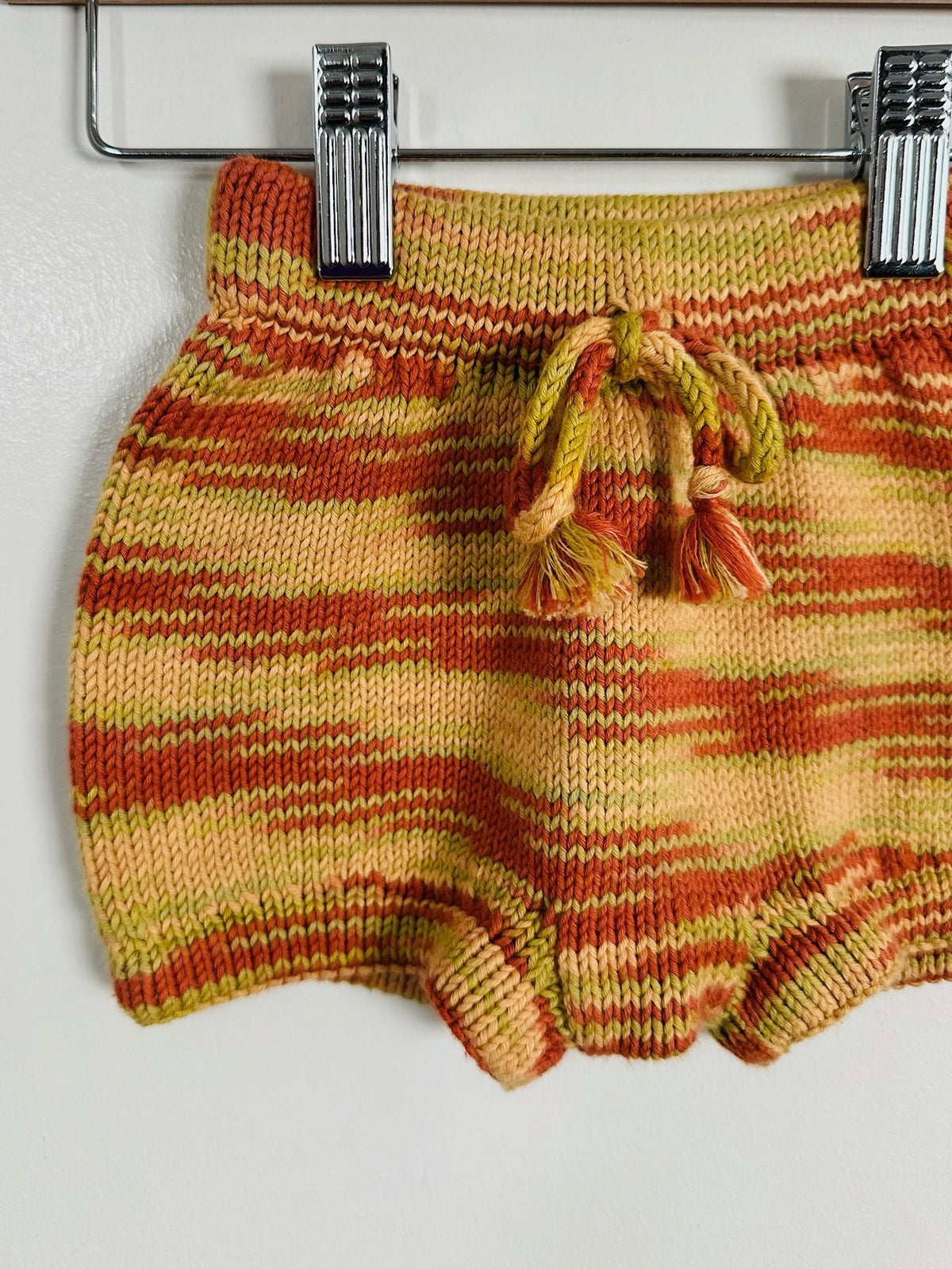 A close-up of a pair of hand-knit baby bloomers in a marled orange, yellow, and green pattern, hanging from a metal clothes hanger against a plain white wall.