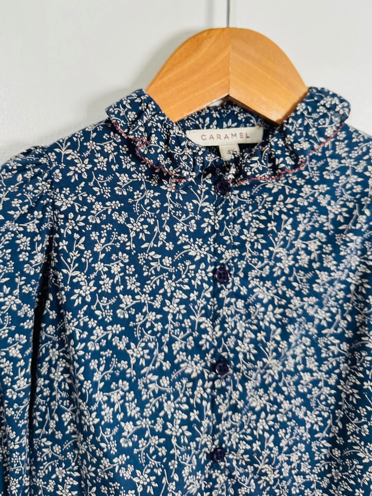 A blue floral blouse with a ruffled collar and button front, hanging on a wooden hanger against a plain wall. The neck label reads Caramel.