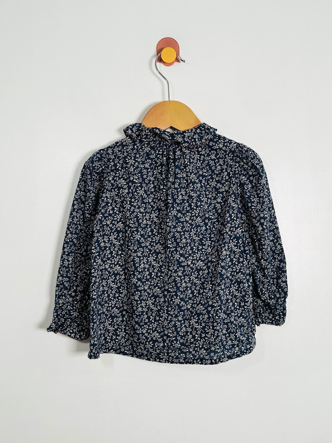 A long-sleeve blouse with a dense small floral print in navy blue and white, hanging on a wooden hanger with a colorful hook against a plain white wall.
