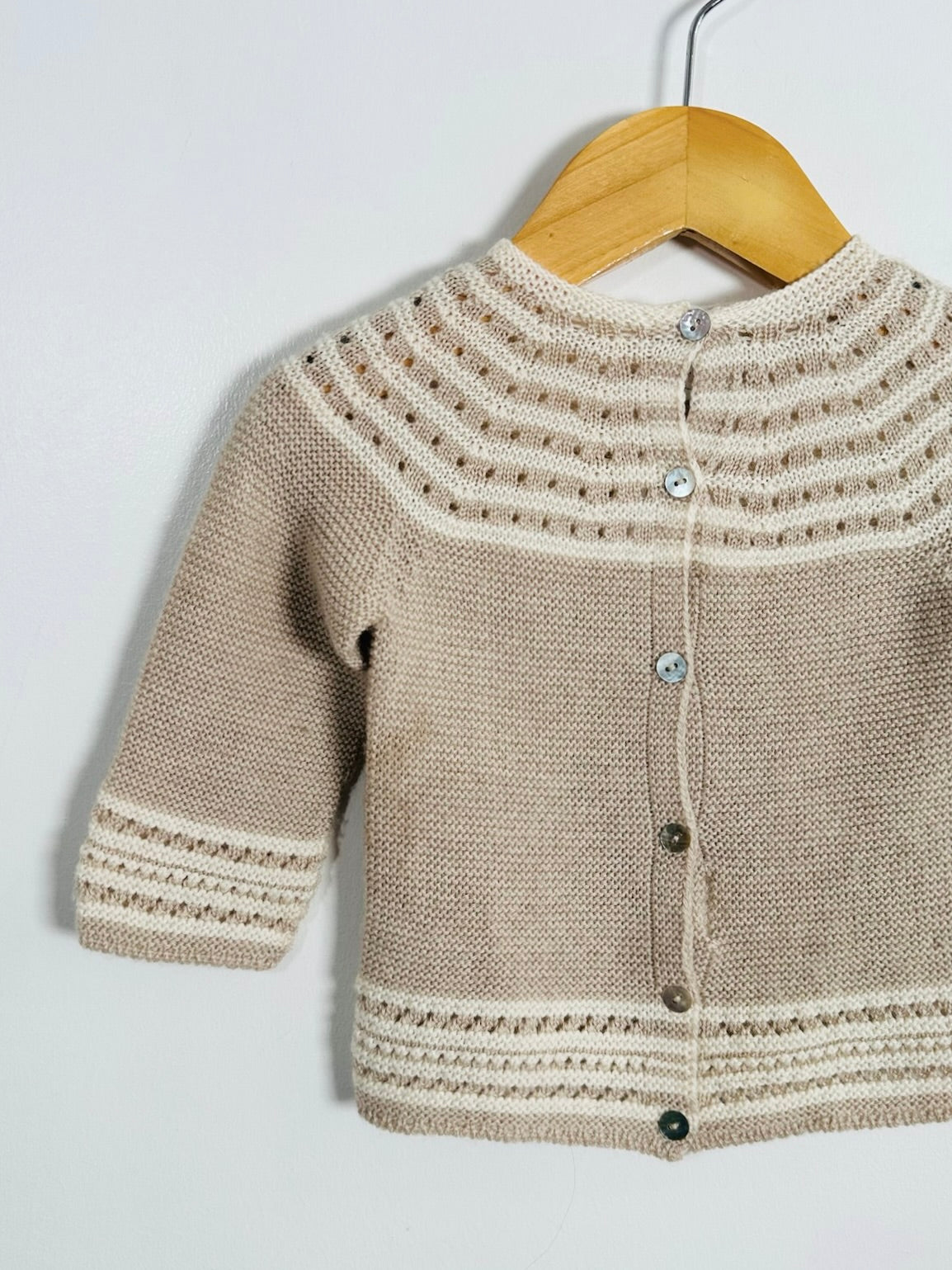 A beige and cream hand-knit baby cardigan sweater with decorative patterns and silver buttons, hanging on a wooden hanger against a plain light gray wall.