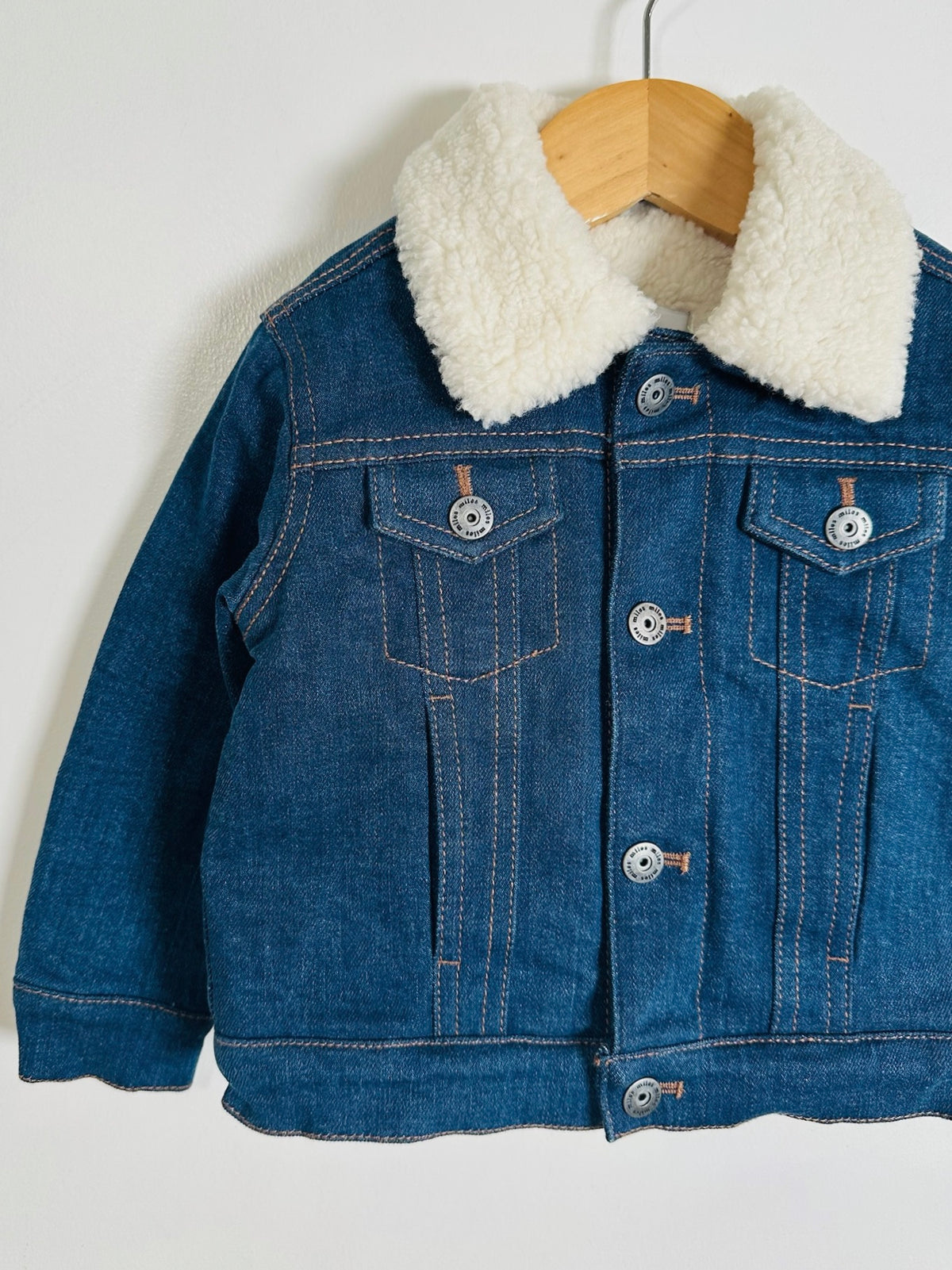 A children's blue denim jacket with a thick cream sherpa fleece collar, hanging on a wooden hanger against a plain white background.
