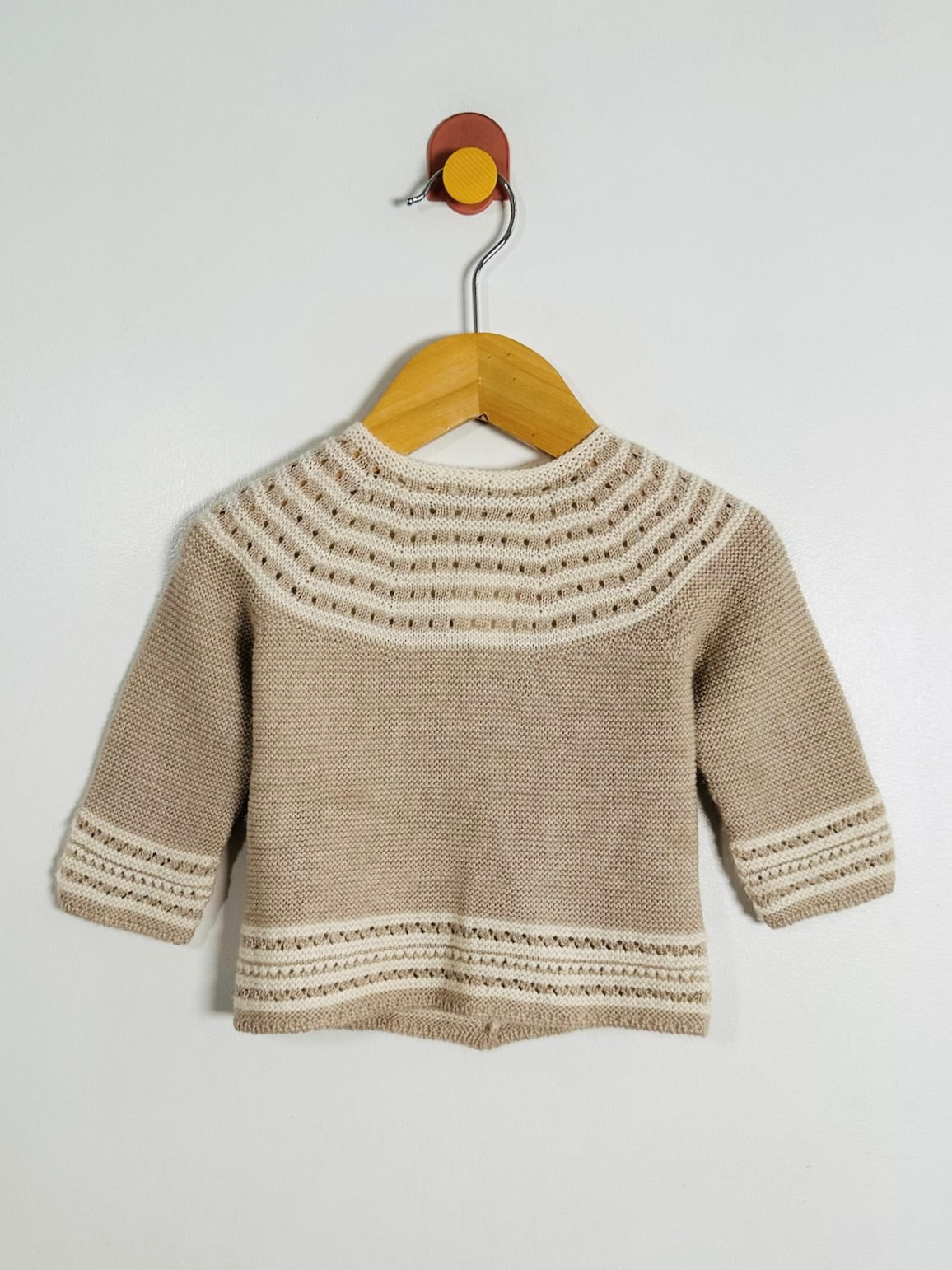 A beige knit baby sweater with cream-colored geometric patterns on the yoke, cuffs, and hem, hanging on a wooden hanger against a plain light gray wall.