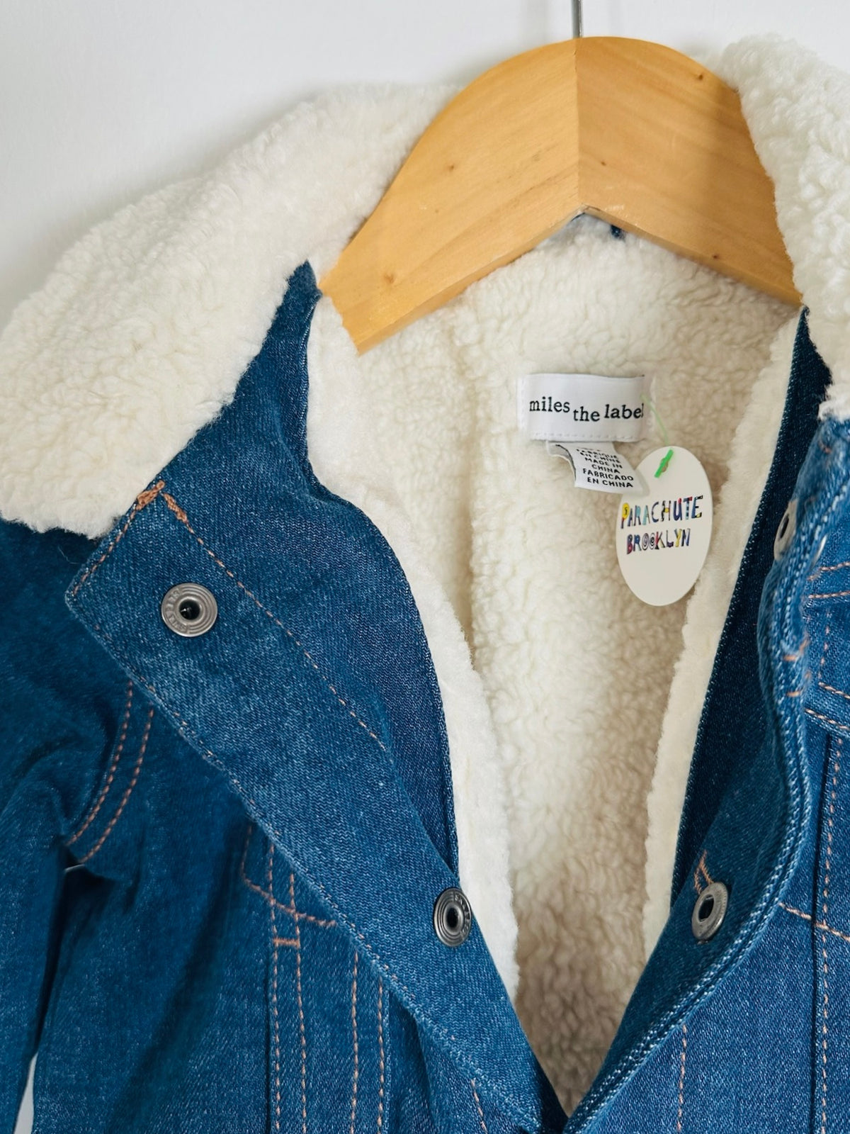 A close-up of a blue denim jacket with a thick white sherpa lining, hanging on a wooden hanger against a white background. The brand label 'miles the label' is visible inside the collar.
