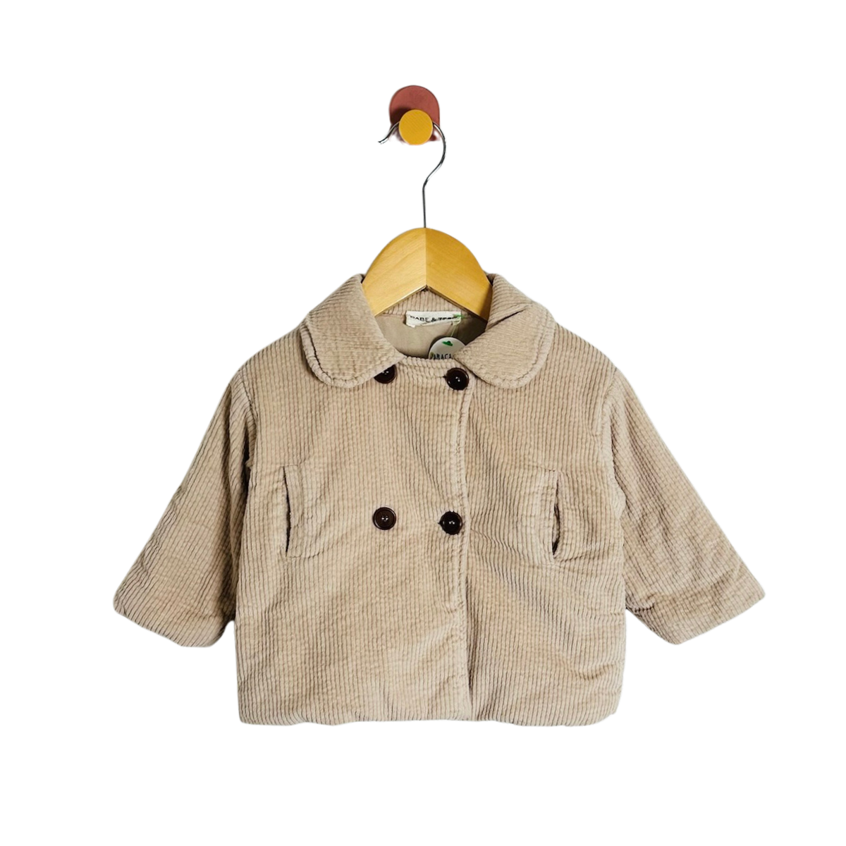 A baby's corduroy jacket in beige, featuring a Peter Pan collar, double-breasted buttons, and front patch pockets, hanging on a wooden hanger against a white background.