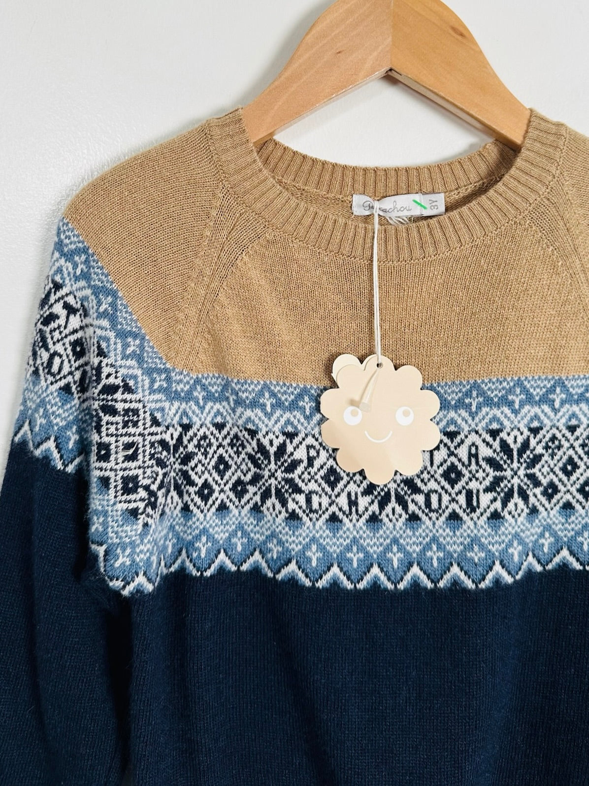 A knit crewneck sweater hanging on a wooden hanger against a plain light wall. The sweater has a tan upper yoke, a light blue and white Fair Isle patterned band across the chest, and a dark navy lower body.