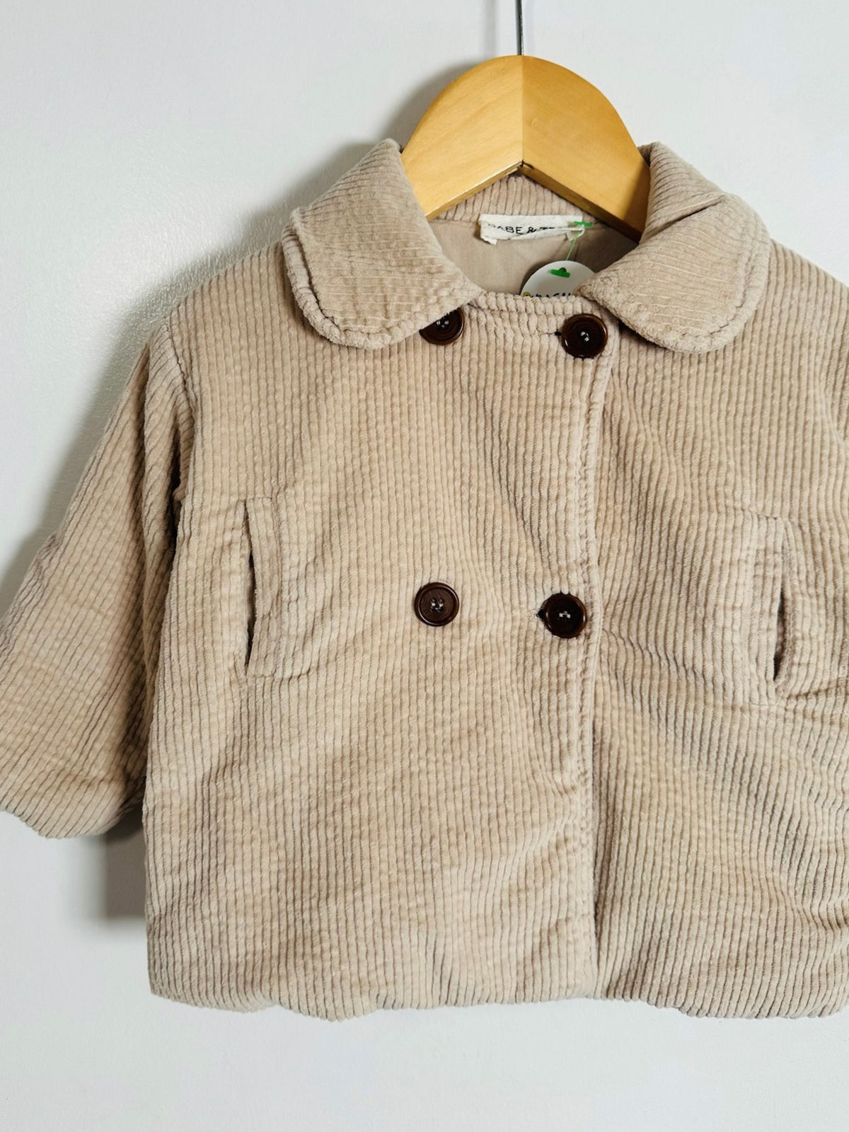 A beige corduroy jacket with a wide collar and dark brown buttons, hanging on a wooden hanger against a plain white wall.