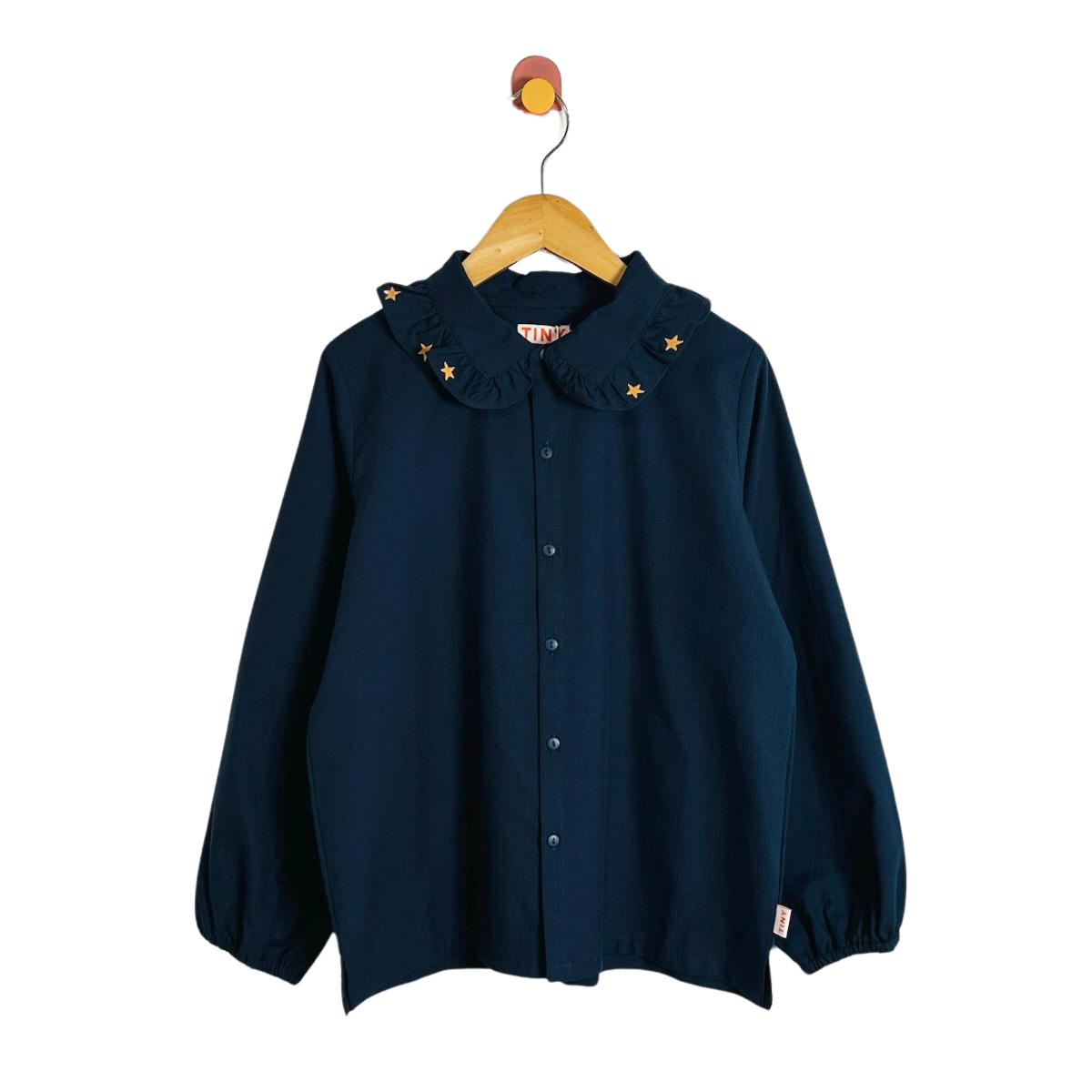 A navy blue long-sleeve button-up shirt with a ruffled Peter Pan collar decorated with small gold star appliqués, hanging on a wooden hanger against a plain white background.