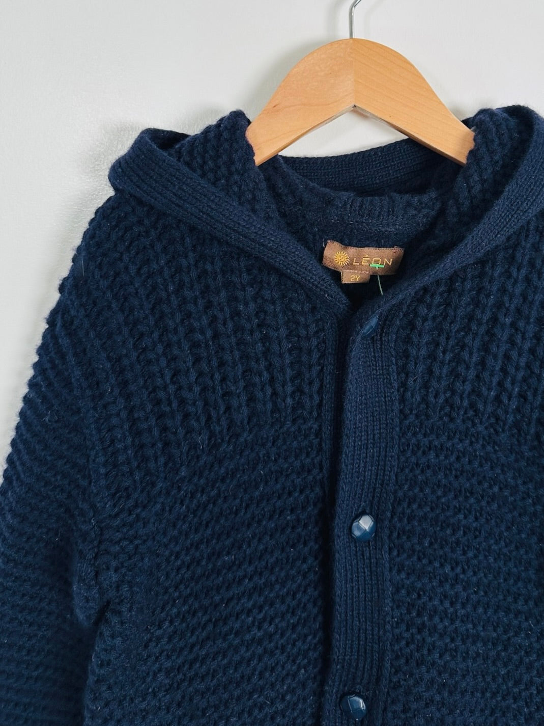 A close-up of a chunky navy blue knit cardigan sweater hanging on a wooden hanger against a plain light background.
