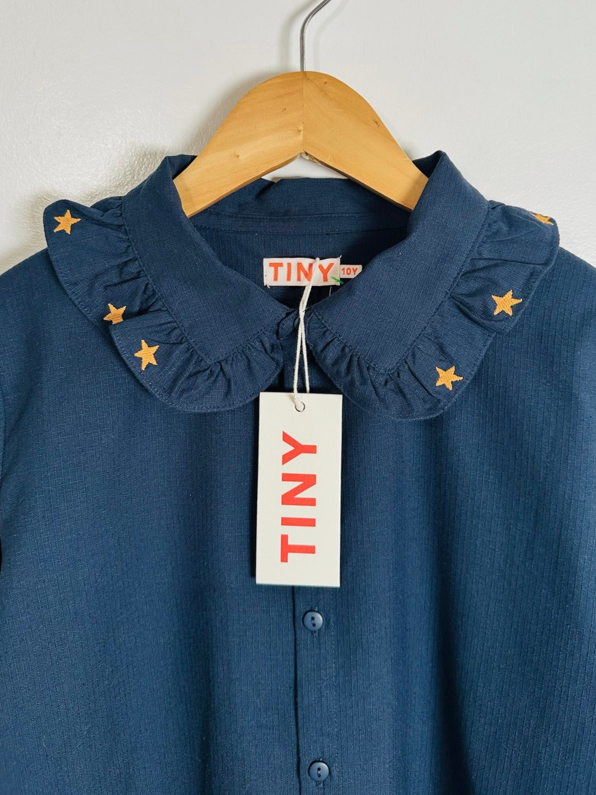 A navy blue button-up shirt with a ruffled Peter Pan collar decorated with small gold stars, hanging on a wooden hanger with a white TINY brand tag.