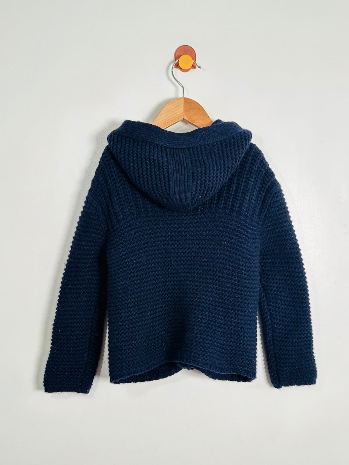 A dark navy blue textured knit hooded sweater hanging on a wooden hanger against a plain light gray wall.