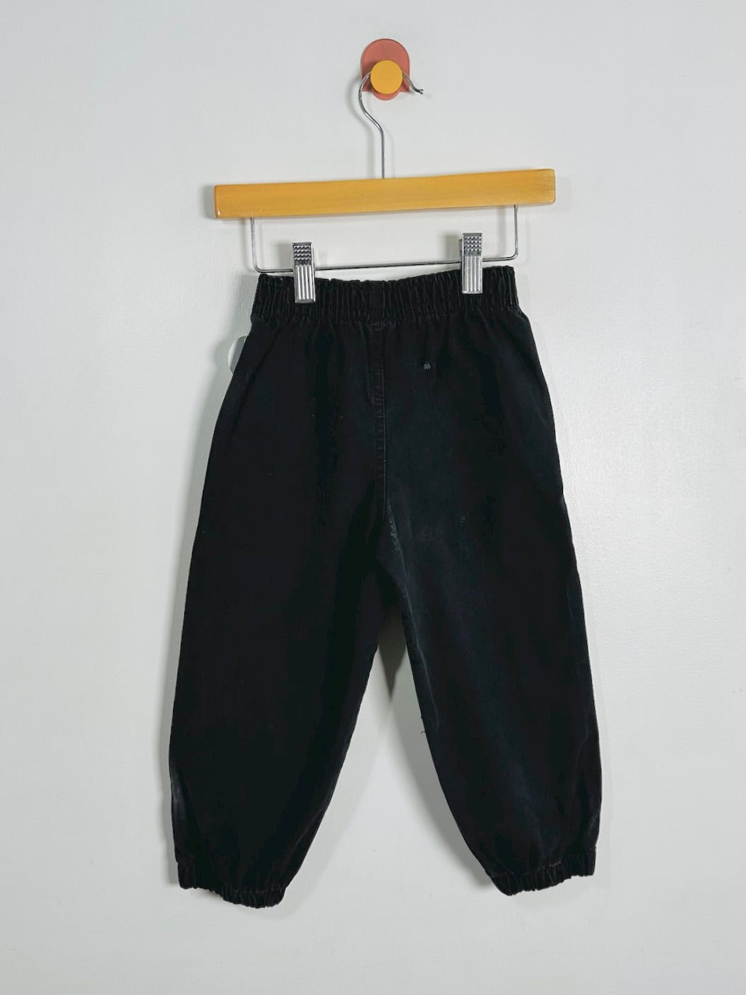 Black wide-leg pants with an elastic waistband and gathered ankle cuffs, hanging from a wooden hanger against a plain light gray wall.