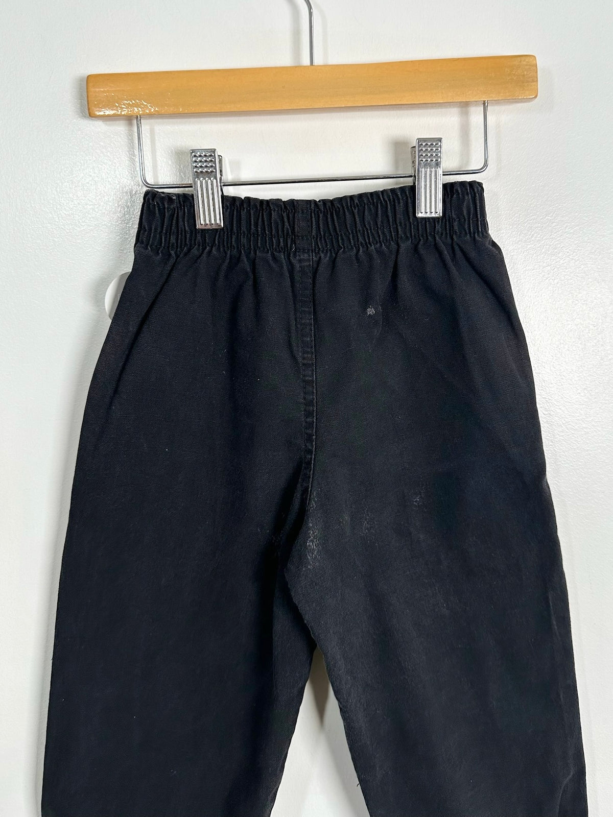 Black wide-leg pants with an elastic waistband, hanging from a wooden hanger with metal clips against a white wall.