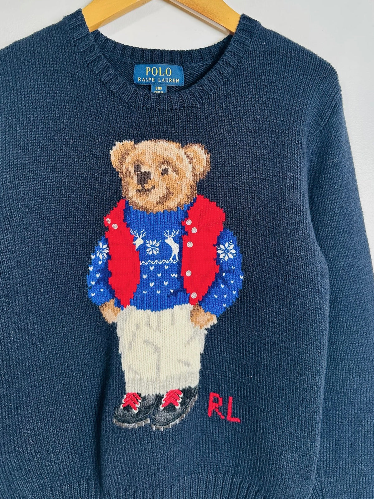 A navy blue knit sweater by Polo Ralph Lauren featuring a knitted teddy bear wearing a blue holiday sweater, red vest, and cream pants, with 'RL' embroidered in red on the lower right.
