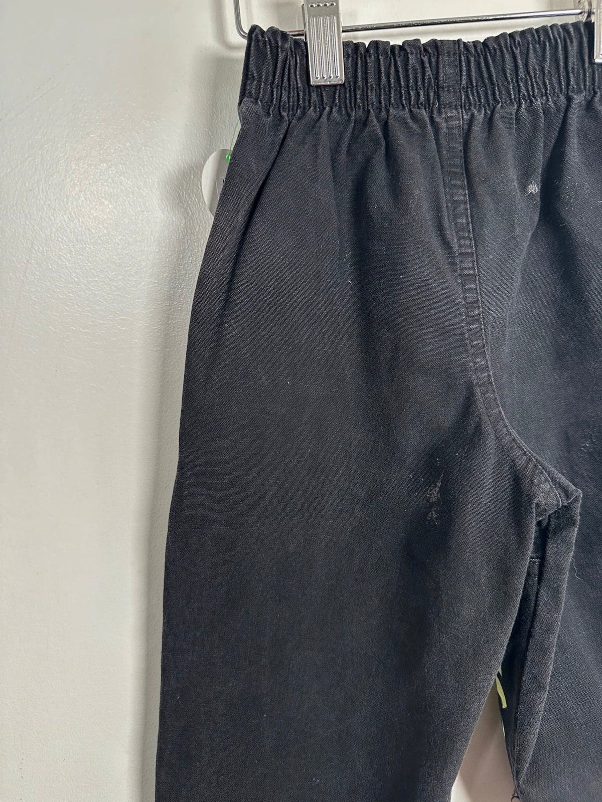 Close-up of a pair of dark charcoal gray or black elastic-waist pants hanging on a metal hanger against a plain off-white wall.
