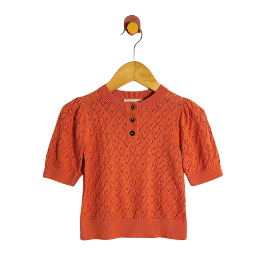 An orange short-sleeve knit top with a textured geometric pattern and three black buttons at the collar, hanging on a wooden hanger against a white background.
