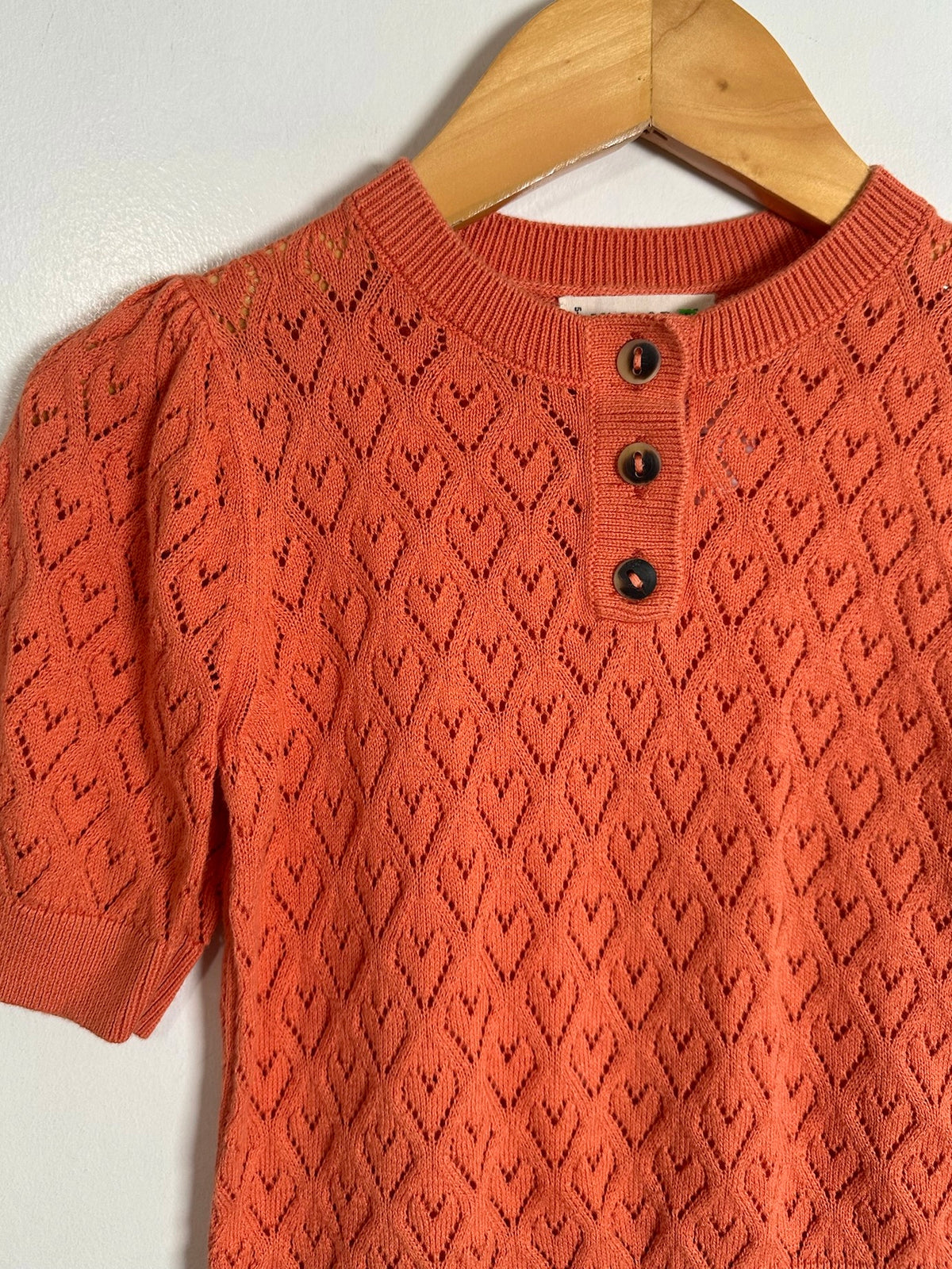 An orange knit short-sleeve top with a three-button henley neckline, hanging on a wooden hanger against a plain light background.