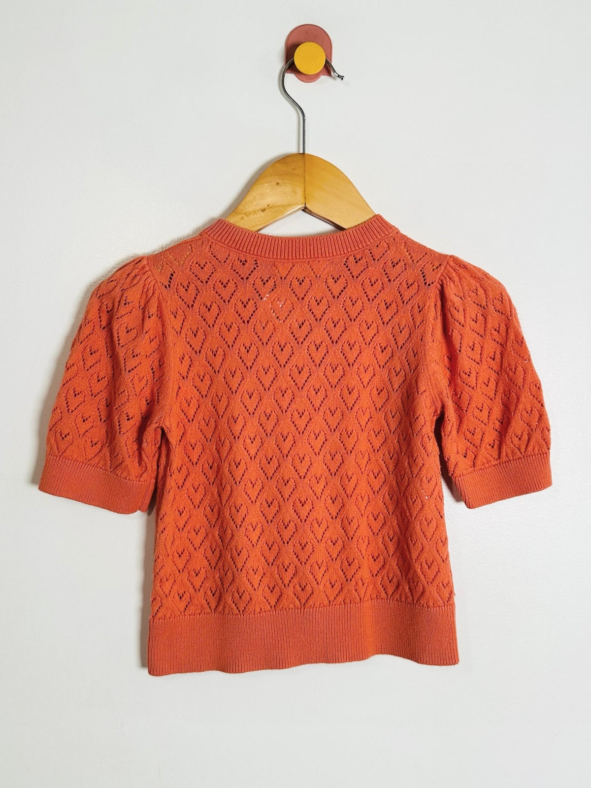An orange short-sleeve knit sweater with a textured geometric pattern, hanging on a wooden hanger against a plain white wall.