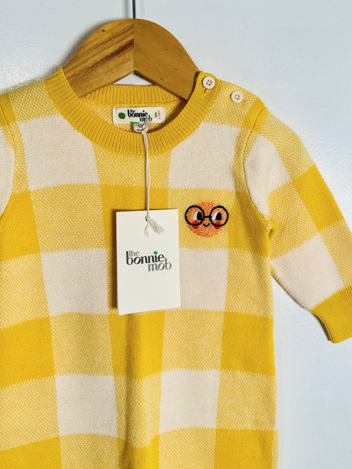 A yellow and cream checkered knit baby sweater with a cartoon face graphic on the chest, hanging on a wooden hanger with a 'the bonnie mob' brand tag.