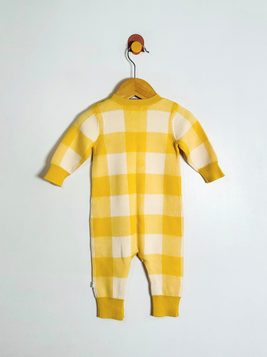 A baby knit one-piece jumpsuit in a yellow and cream gingham check pattern, hanging on a wooden hanger against a plain white wall.