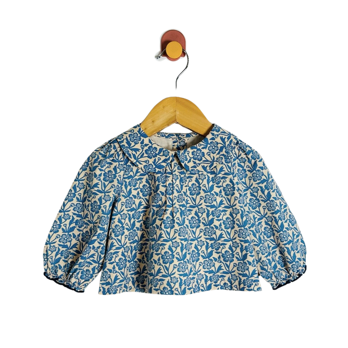 A blue and cream floral print long-sleeve blouse on a wooden hanger, hanging from a small wall hook against a white background.