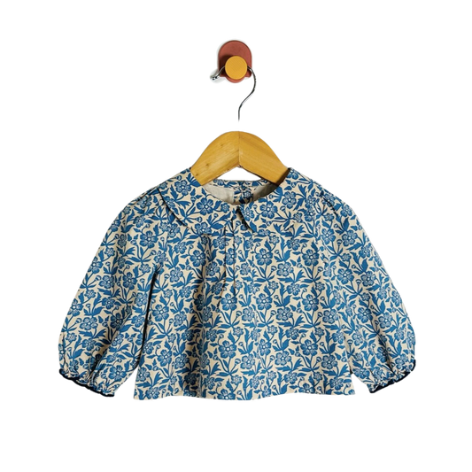 A blue and cream floral print long-sleeve blouse on a wooden hanger, hanging from a small wall hook against a white background.