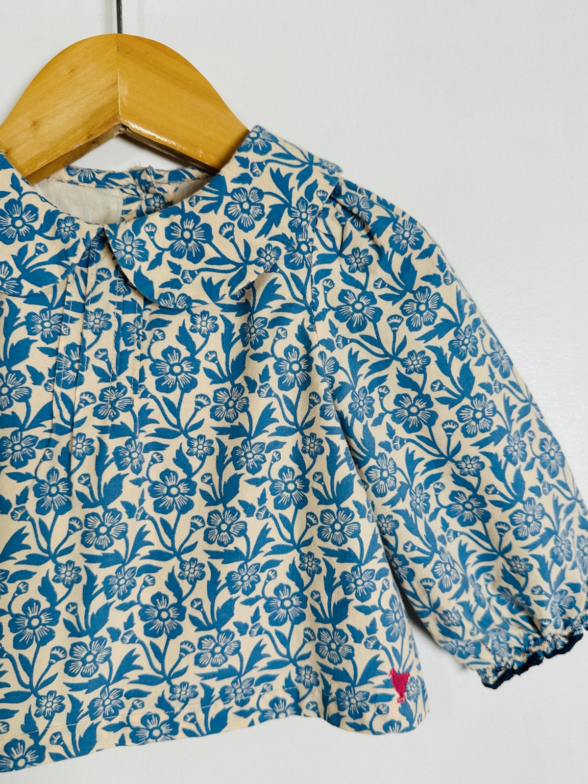 A long-sleeve blouse with a blue floral block-print pattern on a cream-colored fabric, hanging on a wooden hanger against a plain white wall.