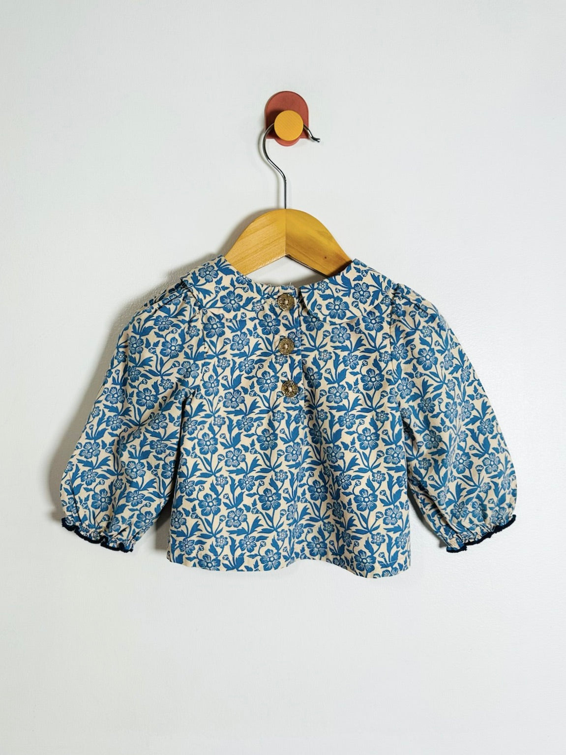 A blue and cream floral print baby jacket with puff sleeves, hanging on a wooden hanger from a wall hook.