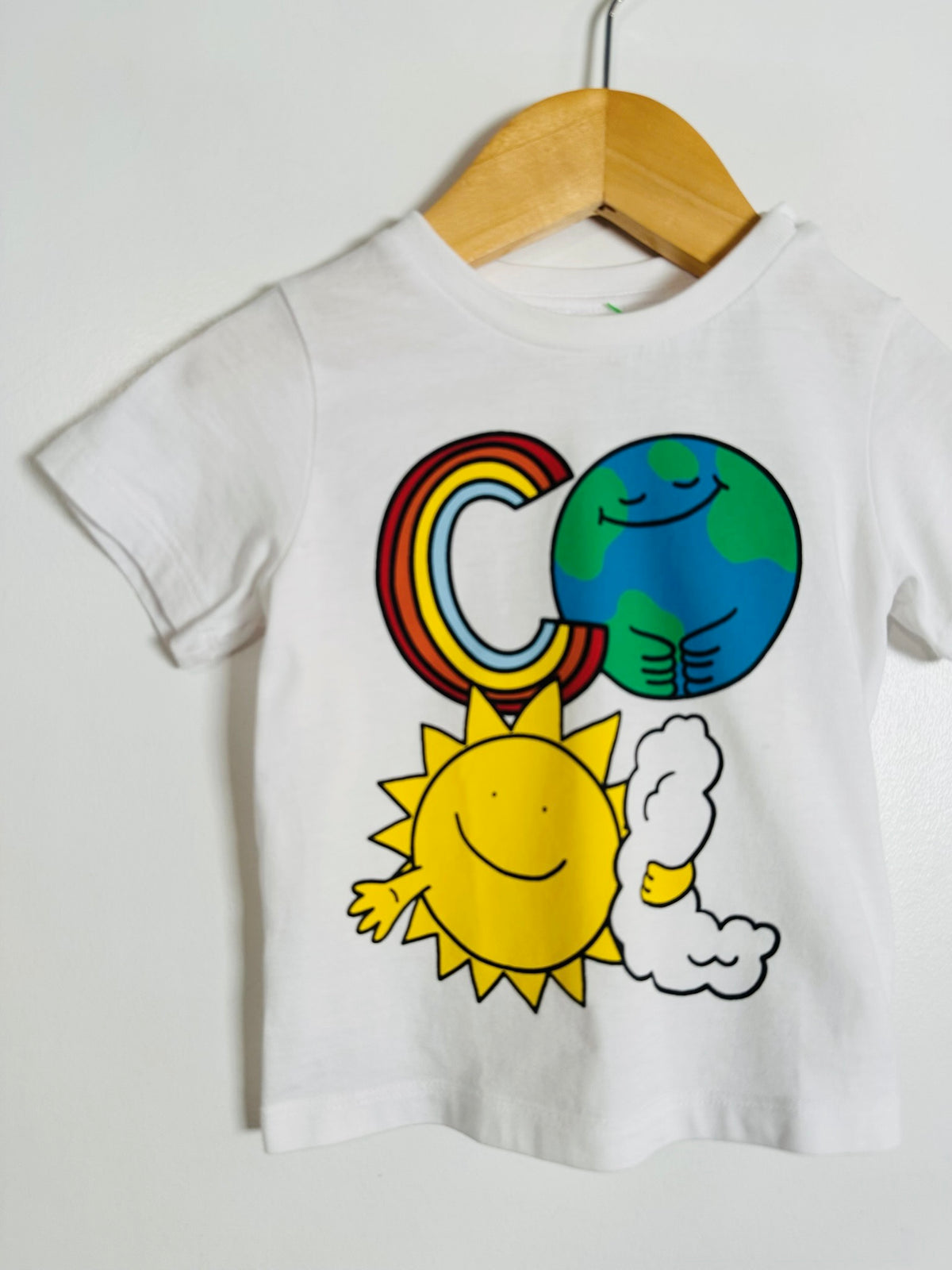 A white short-sleeve kids' t-shirt hanging on a wooden hanger against a plain light wall. The shirt features a colorful graphic of the letter 'C' styled as a rainbow, a blue and green Earth, and a smiling yellow sun with a cloud.