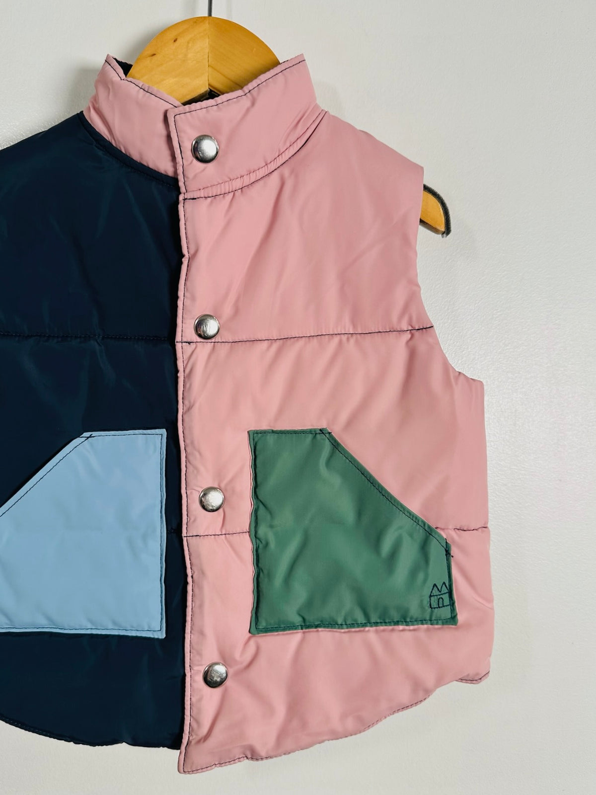 A sleeveless quilted vest hanging on a wooden hanger against a plain light wall. The vest has a split design with a dark navy left side, a pink right side, a light blue triangular patch on the lower left, and a green triangular patch on the lower right.