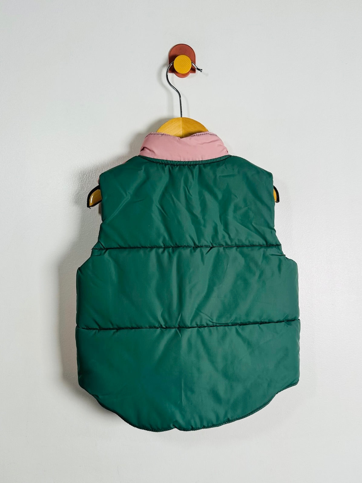 A dark green quilted puffer vest with a pink collar, hanging on a wooden hanger from a wall hook against a plain light gray background.