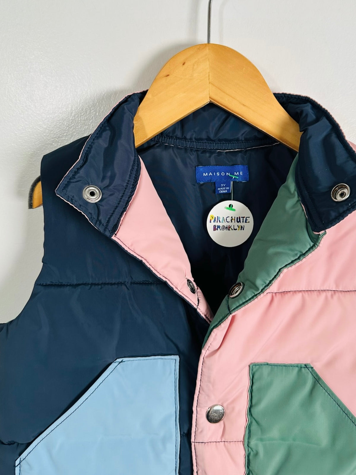 A color-blocked puffer vest by Maison & Pluie hanging on a wooden hanger against a white wall. The vest features panels of navy blue, light blue, pink, and sage green, with silver snap buttons and a high collar.
