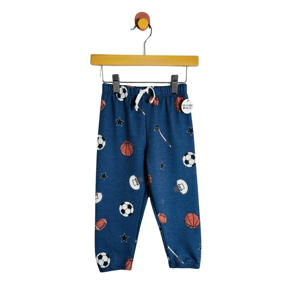 Blue children's jogger pants with a sports-themed print of soccer balls, basketballs, footballs, and stars, hanging on a wooden hanger against a white background.