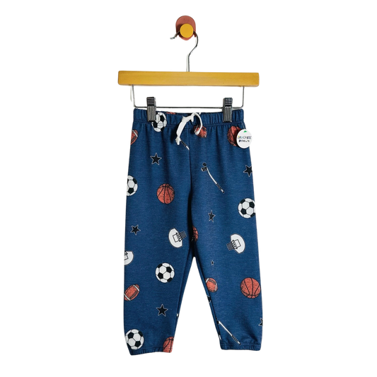 Blue children's jogger pants with a sports-themed print of soccer balls, basketballs, footballs, and stars, hanging on a wooden hanger against a white background.