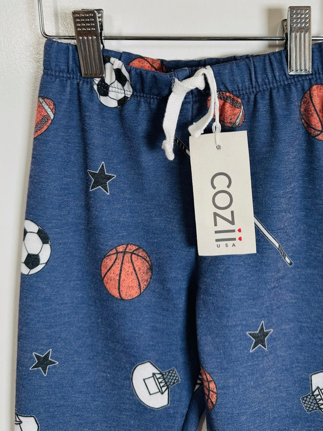 Blue heathered kids' sweatpants with an all-over sports print of basketballs, soccer balls, stars, and a basketball hoop, hanging from a metal rack with a white drawstring and a visible COZIII USA tag.