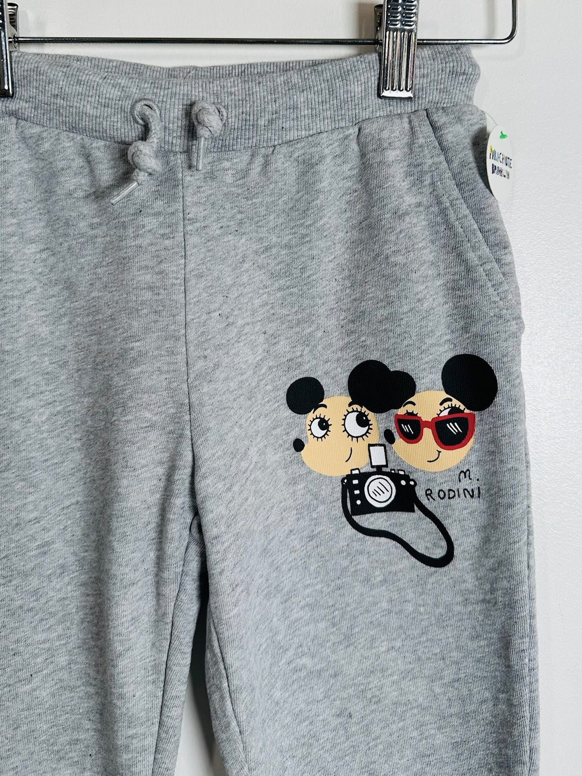 Gray heather sweatpants hanging on a metal hanger, featuring a cartoon graphic of two Mickey Mouse-style characters holding a camera with the text 'M. RODINI' on the front.