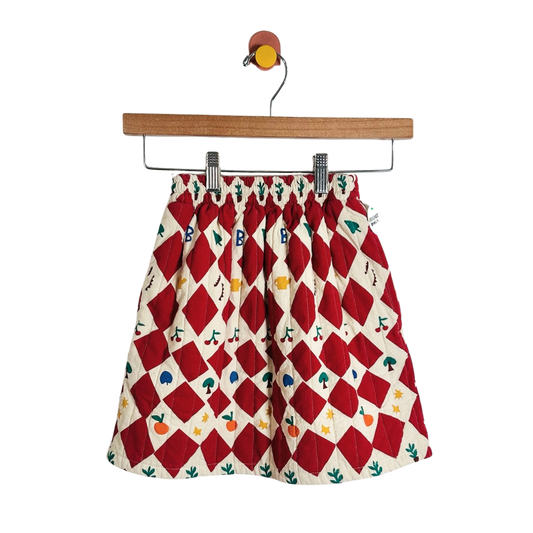 A children's skirt with a red and cream harlequin diamond pattern and small colorful fruit and leaf prints, hanging from a wooden hanger against a plain white background.