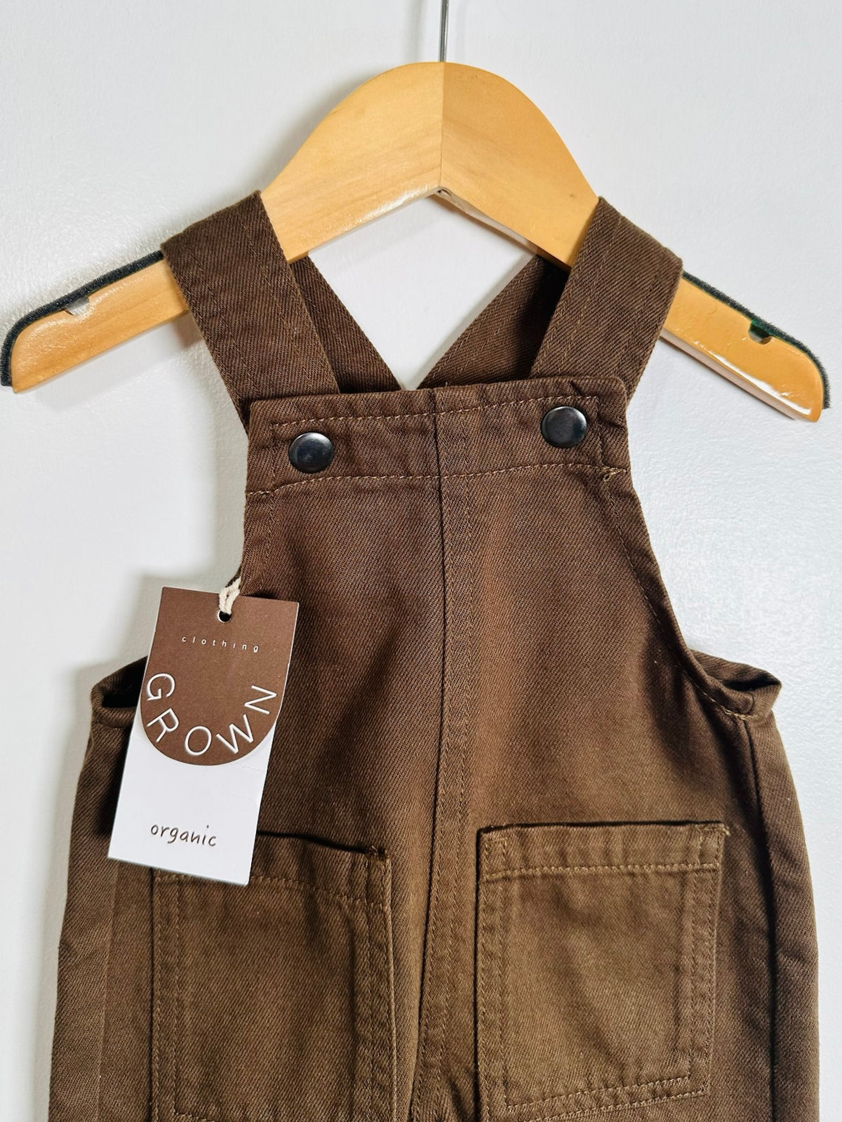 A brown organic cotton overalls hanging on a wooden hanger against a light gray wall. The dress features adjustable shoulder straps with snap buttons, a front button closure, and two front patch pockets.