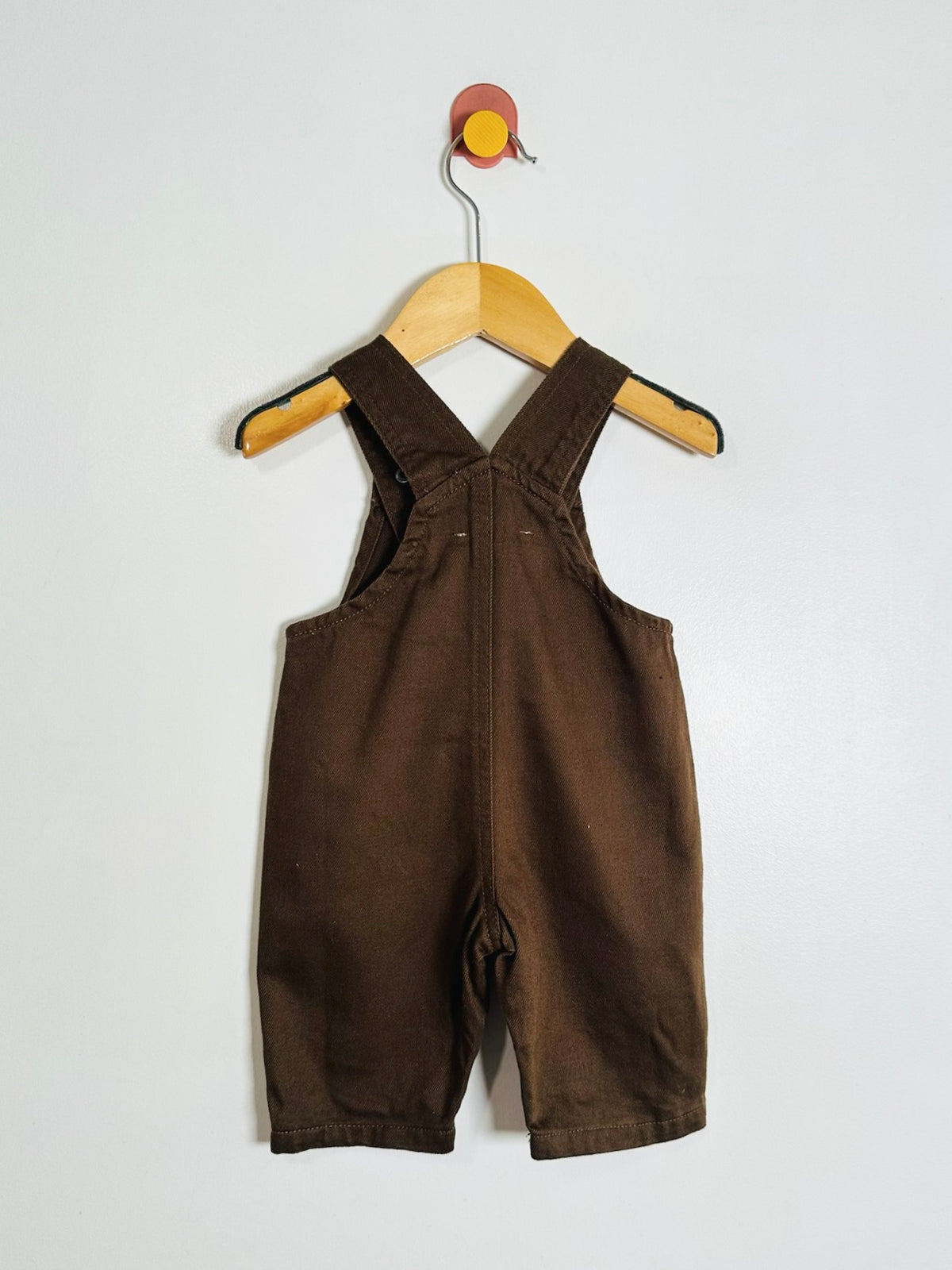 A pair of baby brown corduroy overall shorts hanging on a wooden hanger against a plain white wall.