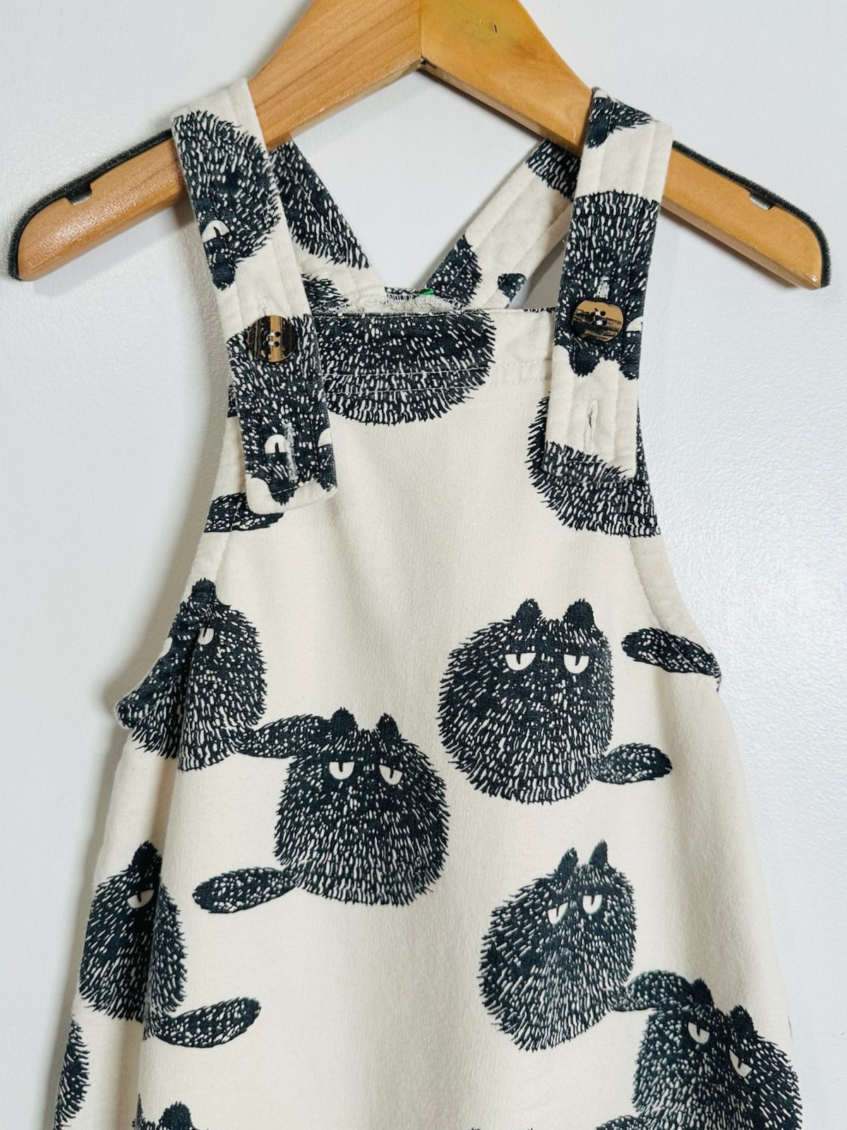 A sleeveless cream-colored dress with a black and white cat print, hanging on a wooden hanger against a plain white wall.