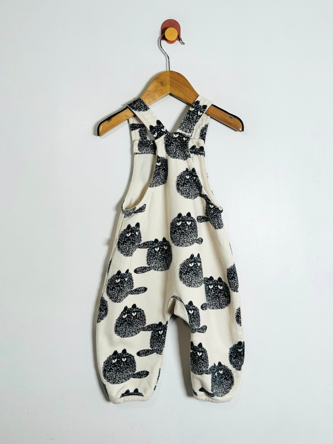 A baby romper in cream fabric with a black cat print pattern, hanging on a wooden hanger against a plain white wall.