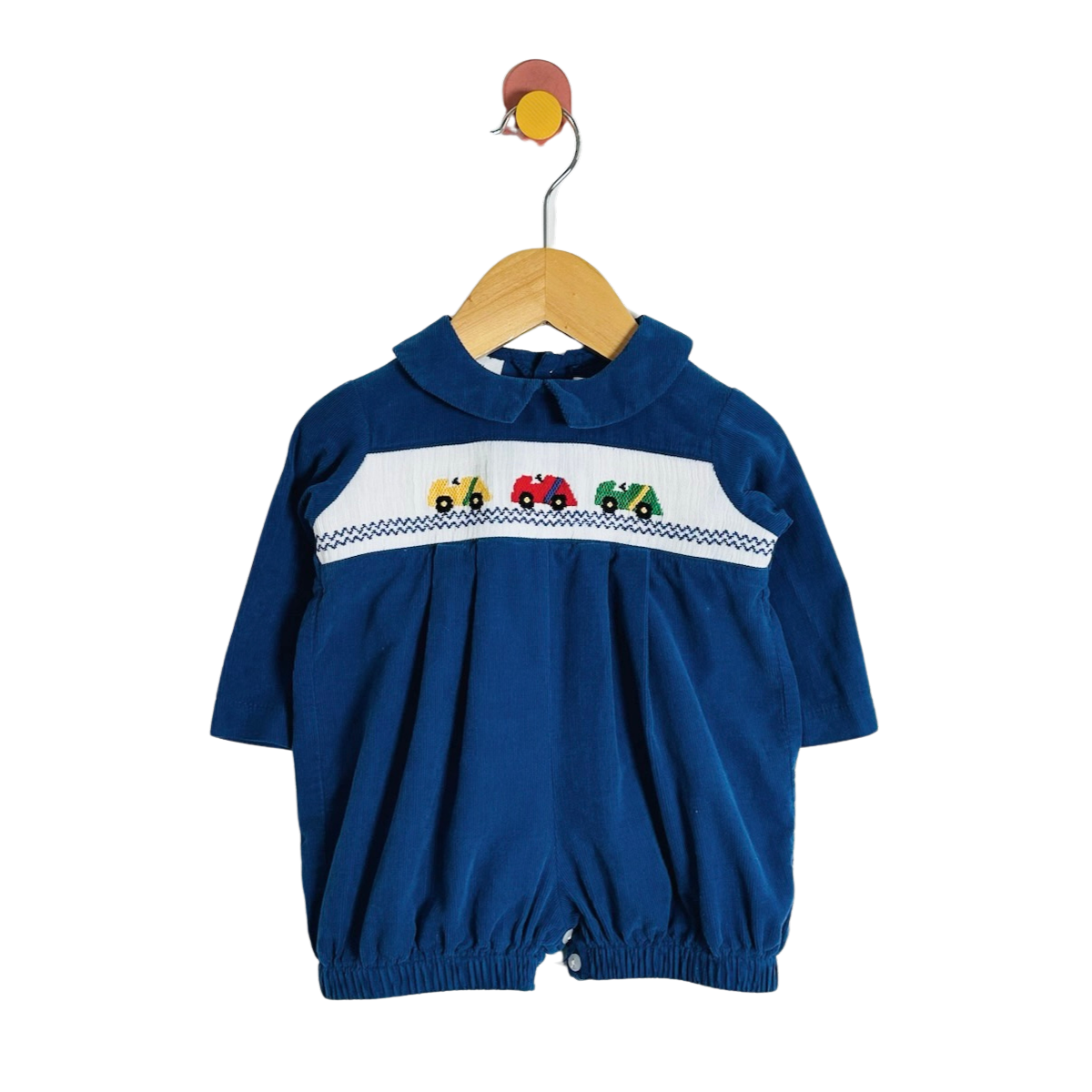 A baby royal blue corduroy jumpsuit with a Peter Pan collar and a decorative chest panel featuring embroidered tractor characters on a white background, hanging on a wooden hanger against a white studio background.