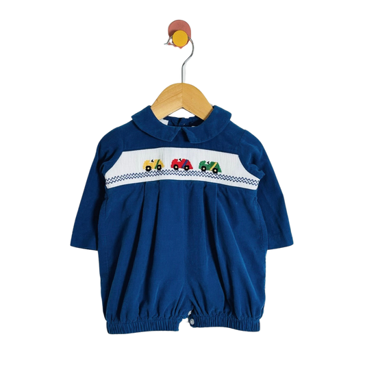 A baby royal blue corduroy jumpsuit with a Peter Pan collar and a decorative chest panel featuring embroidered tractor characters on a white background, hanging on a wooden hanger against a white studio background.