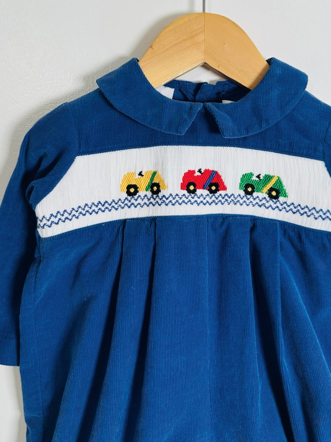 A blue corduroy baby romper with a white smocked chest panel featuring three embroidered toy vehicles (yellow, red, and green), hanging on a wooden hanger against a white wall.