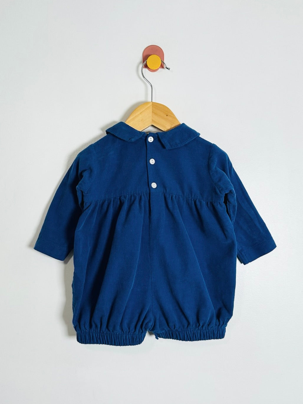 A dark blue corduroy baby romper with a collar and three front buttons, hanging on a wooden hanger against a plain light gray wall.