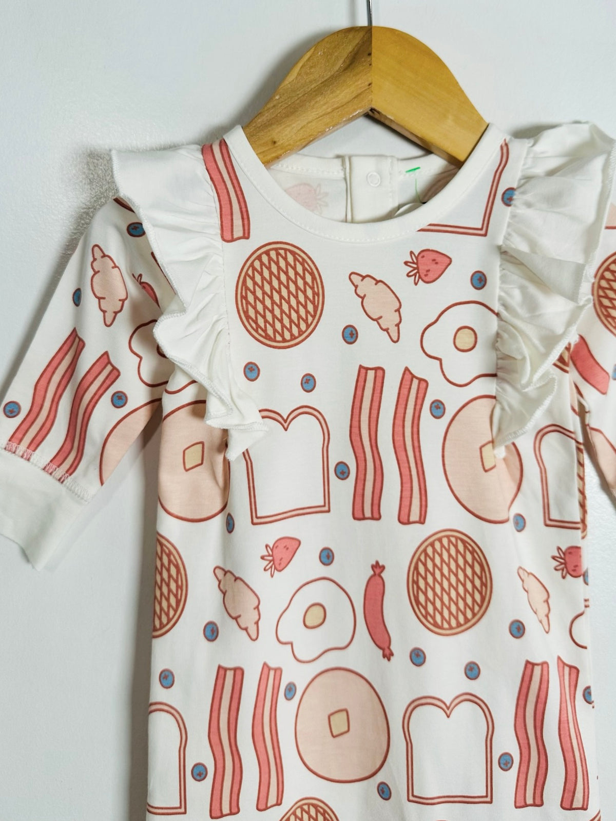 A white long-sleeve baby dress with ruffled shoulders, featuring a playful print of breakfast food illustrations like eggs, bacon, toast, and strawberries in pink and red tones, hanging on a wooden hanger against a white wall.